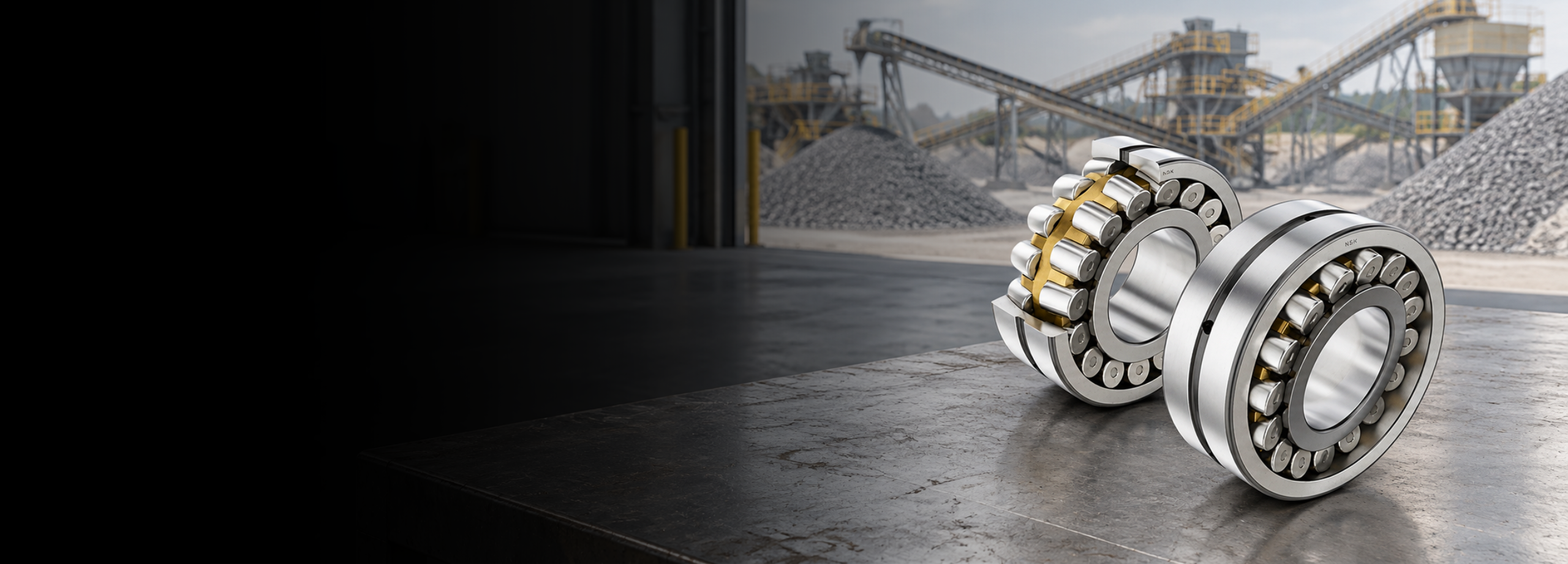 Two NSK spherical roller bearings with brass cages, one sectioned to show the roller rows, on a ledge overlooking an aggregate crushing and screening plant